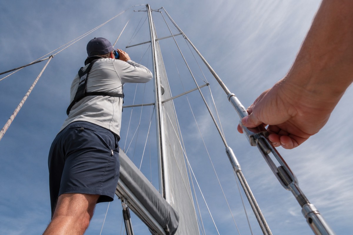 A sailor sighting up a mast while another checks a turnbuckle at deck level